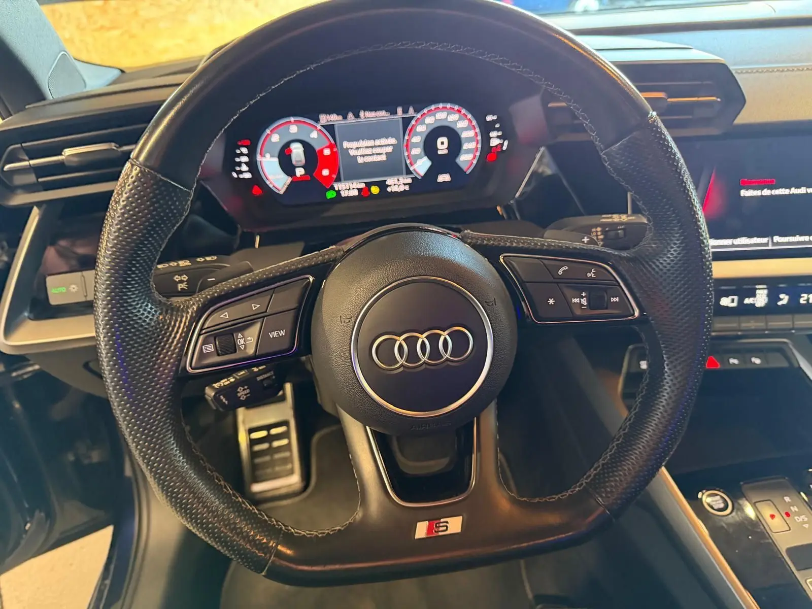 Vue rapprochée du volant cuir perforé noir avec logo Audi et badge S, tableau de bord digital et console centrale visibles.