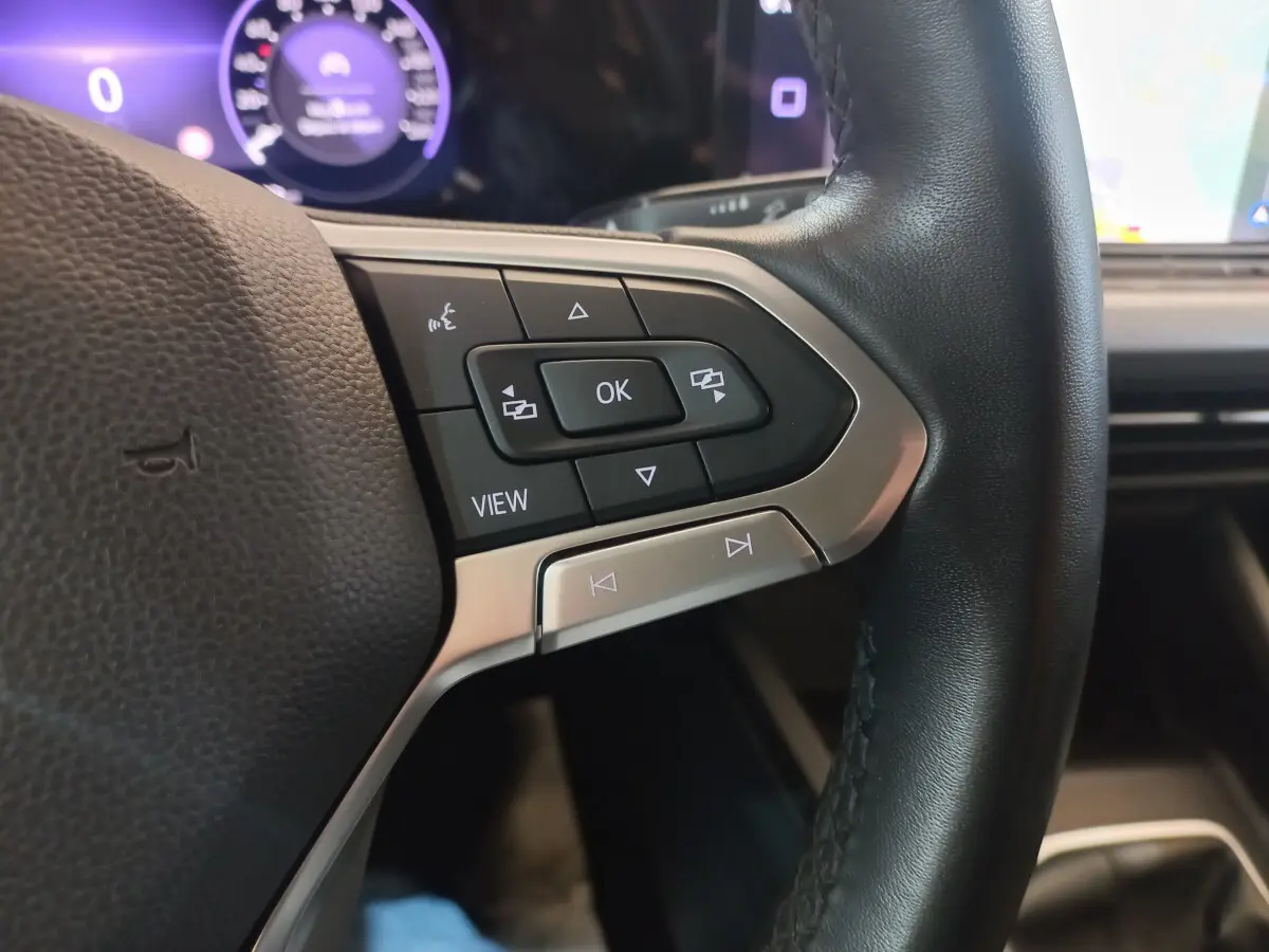 Gros plan sur les commandes multifonctions côté gauche du volant cuir de la Volkswagen Golf 2022 avec affichage digital violet en arrière-plan.