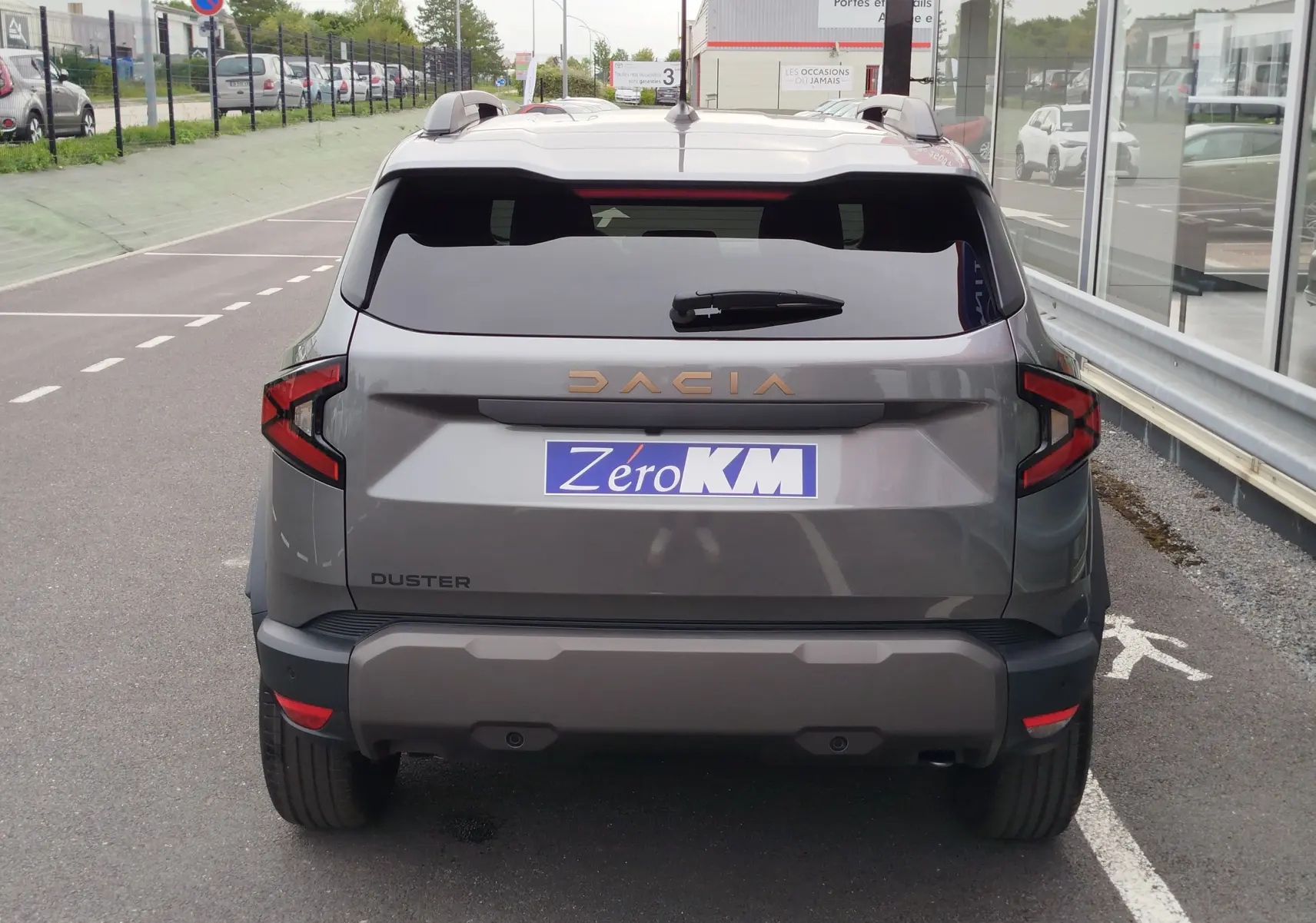 Vue arrière d'un Dacia Duster gris schiste 2025 avec logo doré et plaque Zéro KM visible sur un parking.