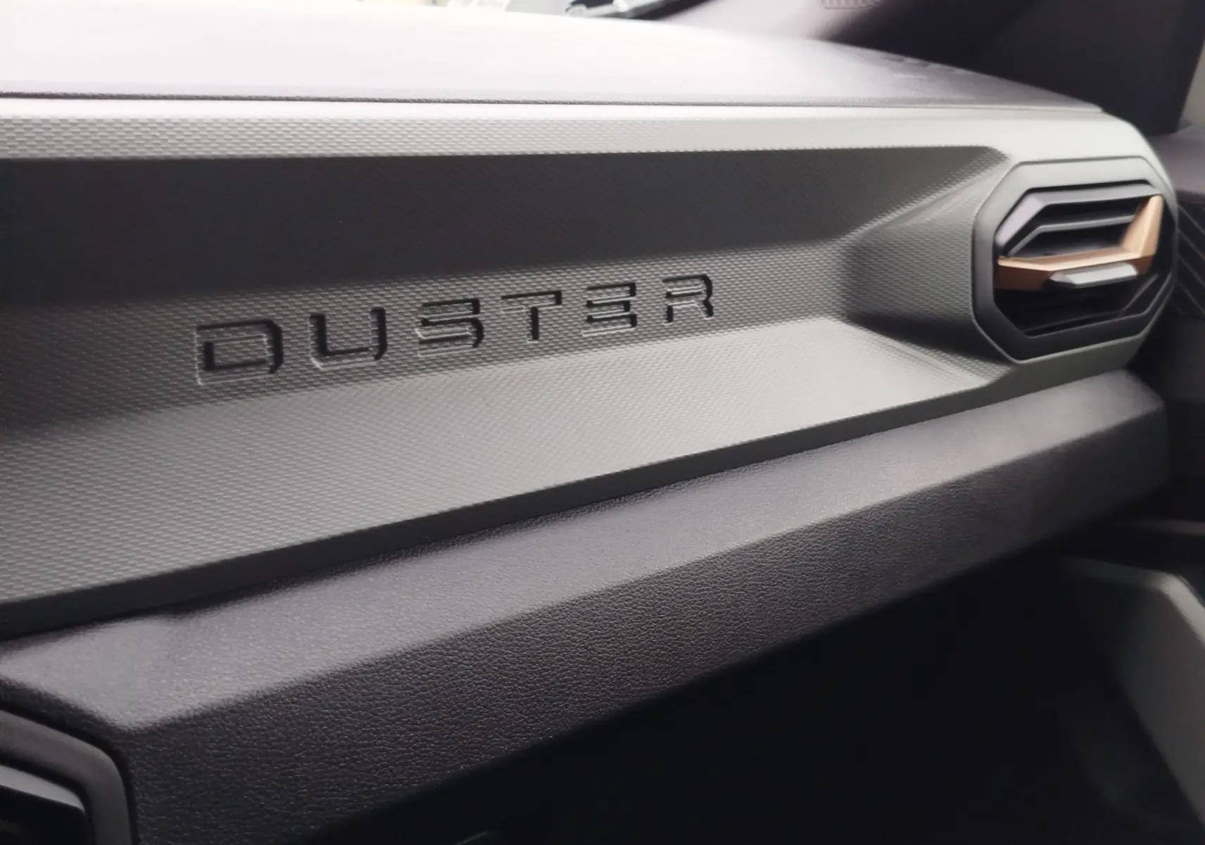 Détail intérieur du tableau de bord gris avec inscription DUSTER et sortie d'air au design hexagonal.