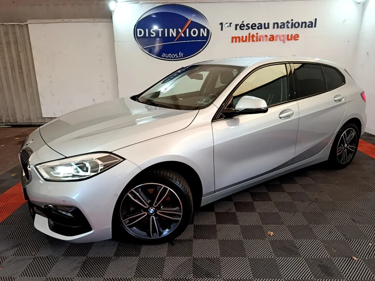 BMW Série 1 gris clair vue 3/4 avant droit, phares LED allumés et jantes sport noires en showroom