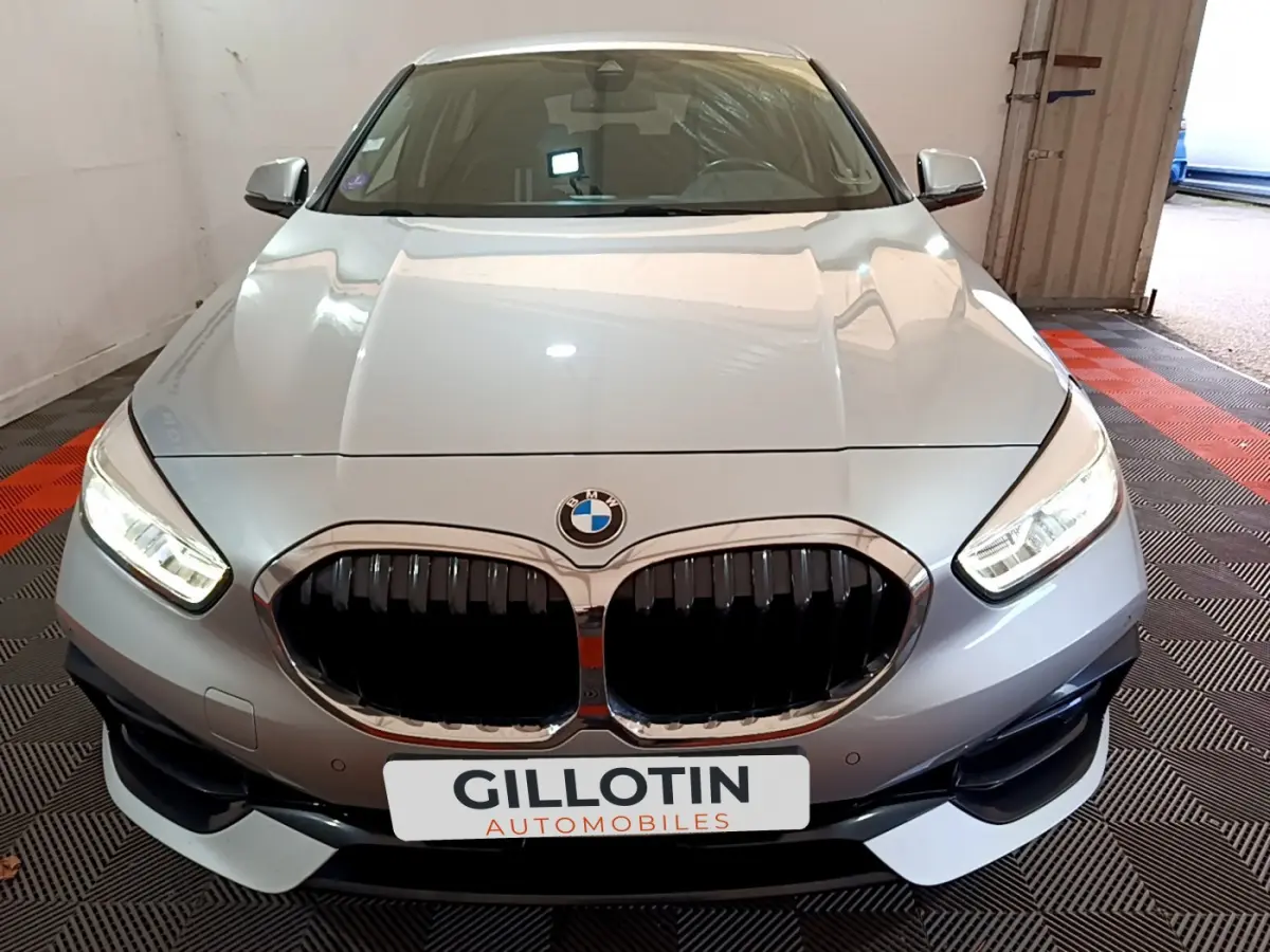 Vue avant d'une BMW Série 1 gris clair avec calandre noire et phares LED allumés en intérieur showroom.