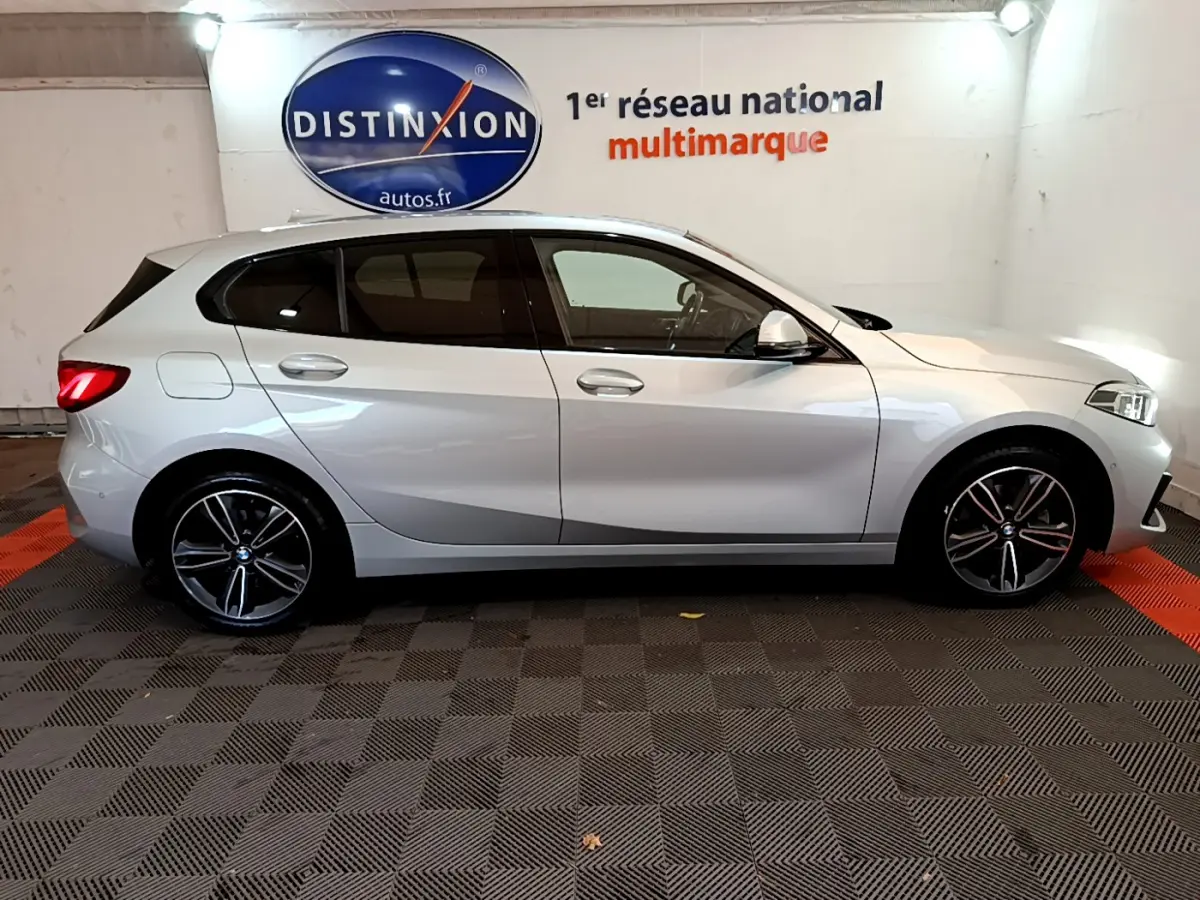 BMW Série 1 gris clair vue de profil côté gauche, avec jantes alliage et feux arrière allumés dans un showroom.