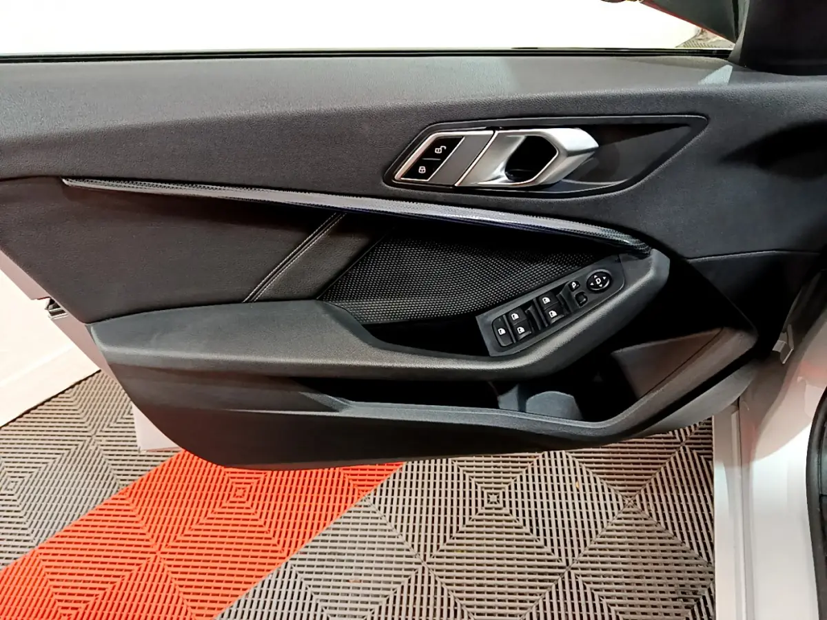 Porte côté conducteur ouverte d'une BMW Série 1 gris clair, montrant les commandes électriques des vitres et la poignée intérieure noire.