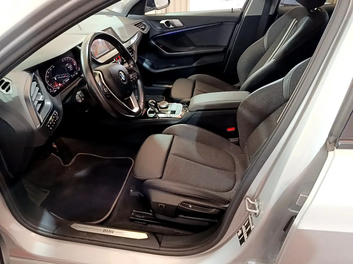 Intérieur avant droit d'une BMW Série 1 gris clair, montrant sièges tissu, volant cuir multifonctions et tableau de bord digital.