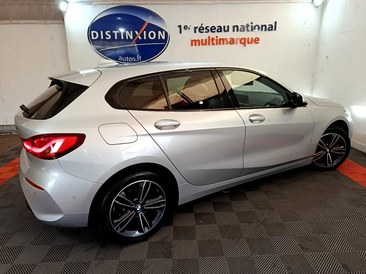 BMW Série 1 gris clair vue de profil côté droit, avec jantes alliage et feux arrière allumés en intérieur showroom.