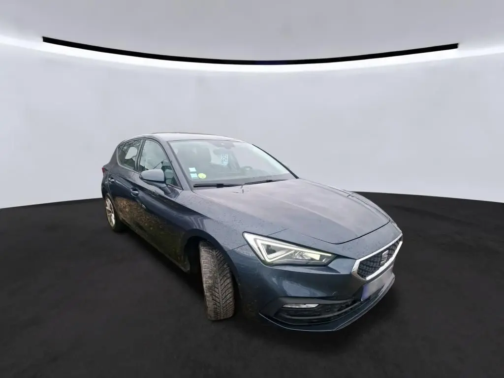 SEAT Leon gris 3/4 avant droit, phares allumés, roues légèrement tournées, sur fond neutre intérieur.