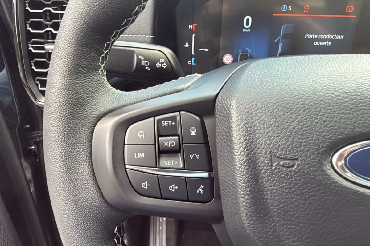 Gros plan sur le volant cuir multifonctions noir du Ford Ranger 2.3 PHEV avec commandes de régulateur et audio, tableau de bord digital visible.