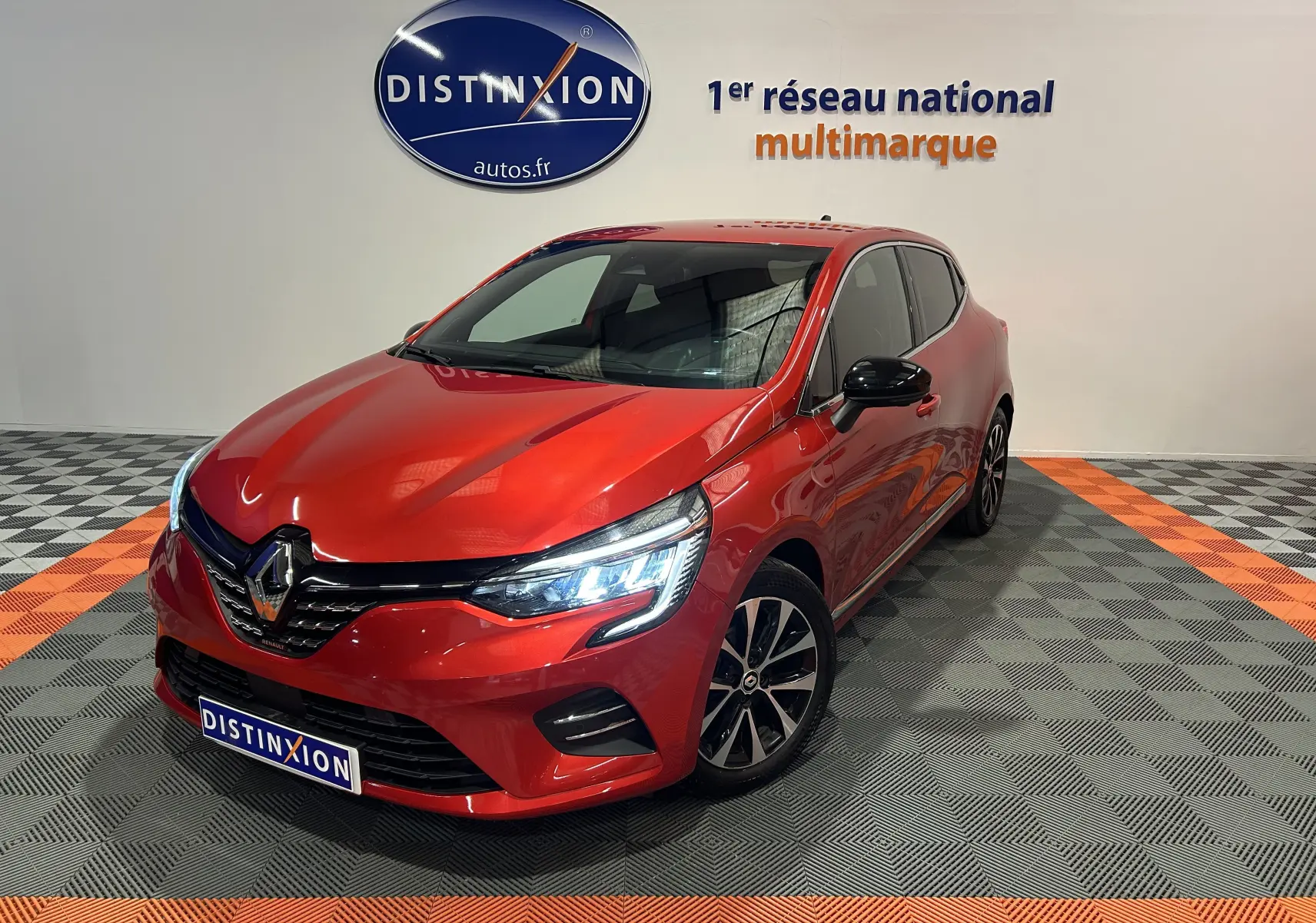 Renault Clio V TCe 90 rouge vue 3/4 avant droit avec phares allumés dans un showroom multimarque