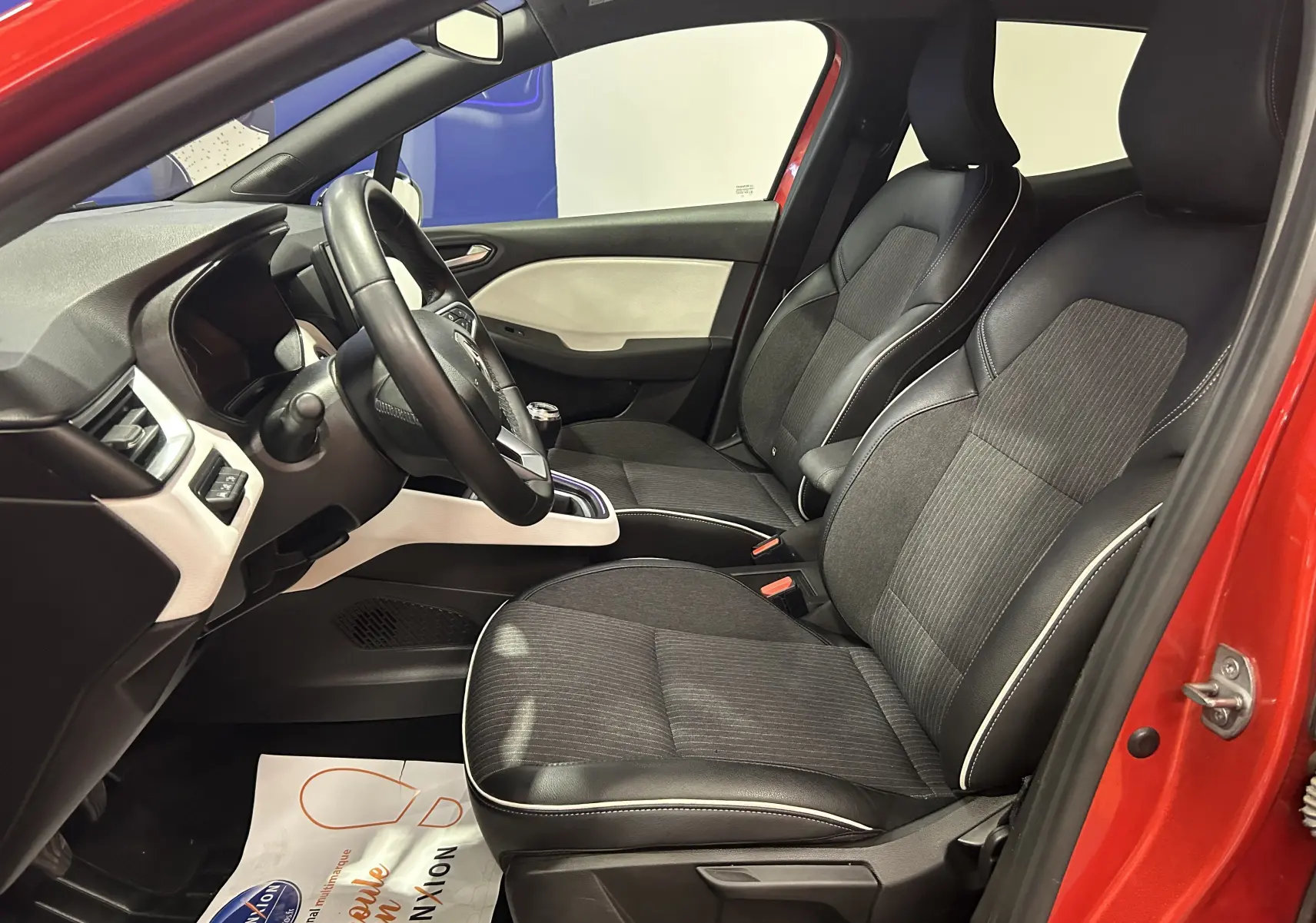 Intérieur avant gauche d'une Renault Clio V rouge 2023, sièges tissu et cuir avec surpiqûres blanches visibles.