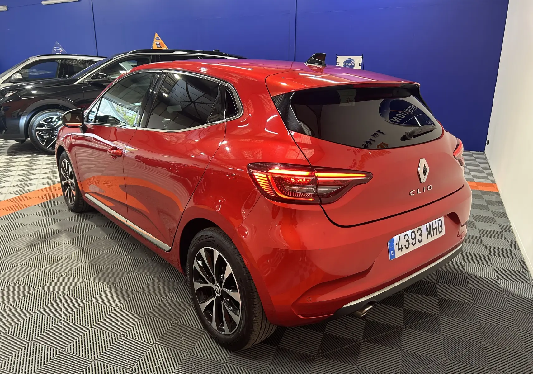 Vue 3/4 arrière droite d'une Renault Clio V rouge 2023 avec feux allumés dans un showroom intérieur.
