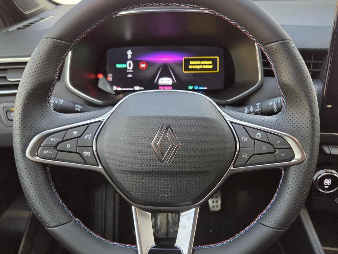 Vue rapprochée du volant perforé noir avec surpiqûres tricolores et logo Renault au centre du tableau de bord numérique.