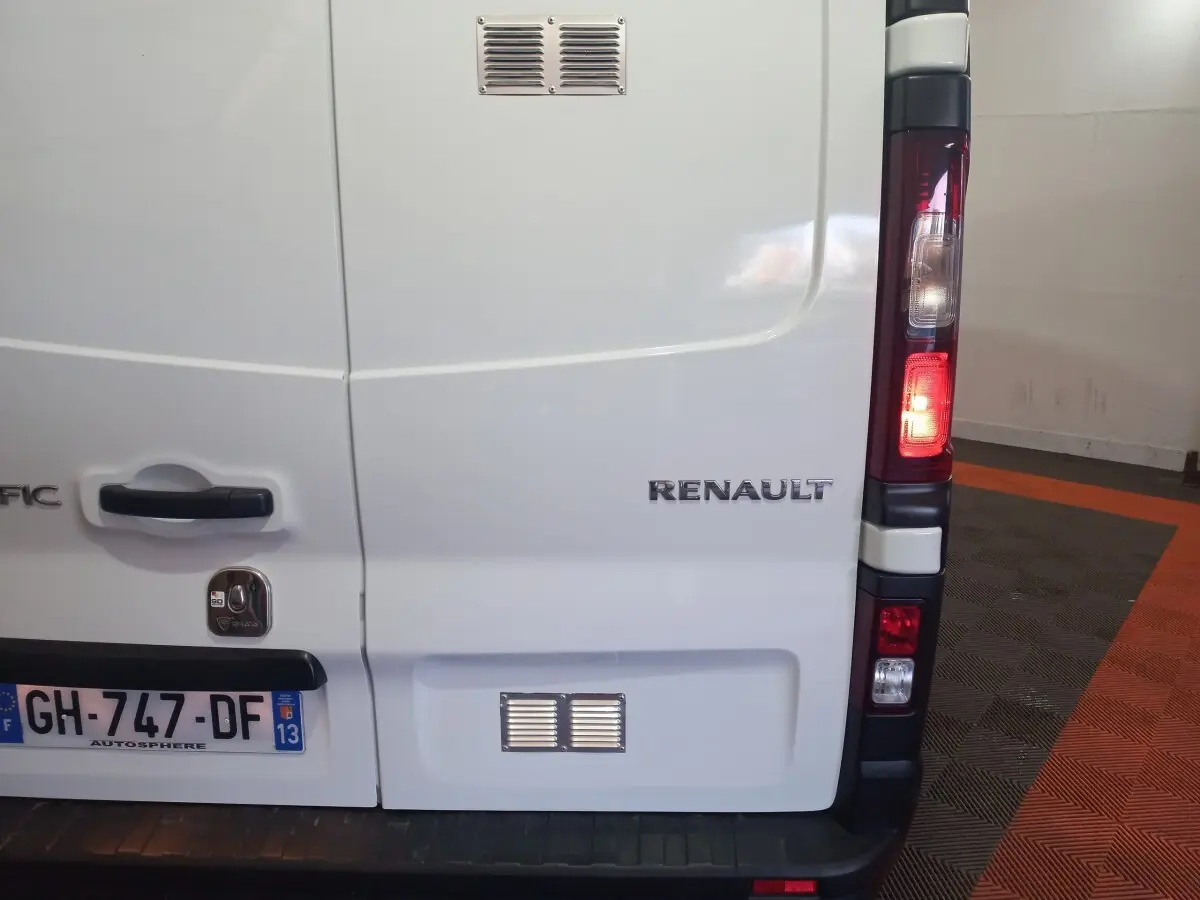 Vue arrière droite du Renault Trafic Fourgon blanc avec feux arrière allumés et logo visible sur la porte.