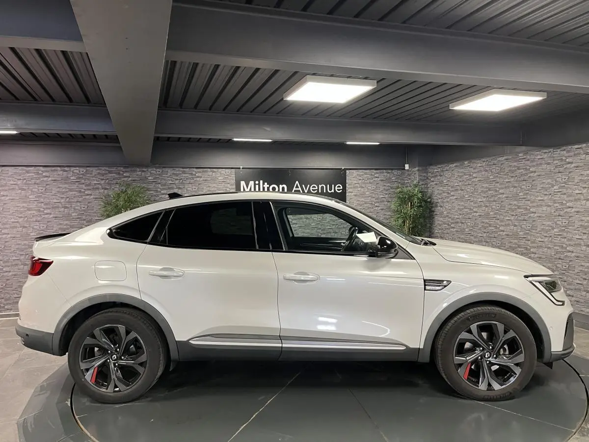 Profil côté gauche du Renault Arkana blanc perle 2022 avec jantes noires et toit noir brillant en intérieur showroom.