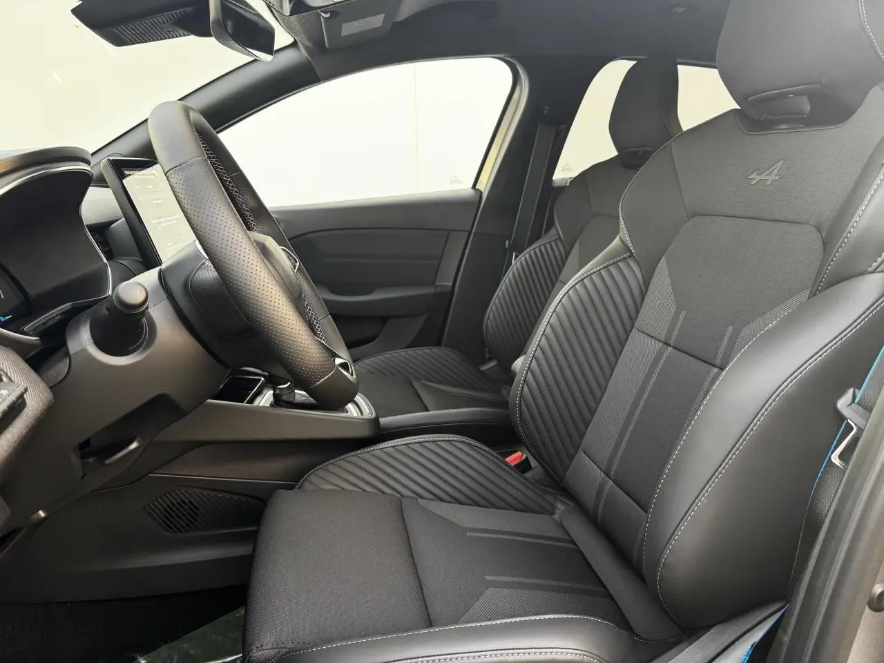 Intérieur avant droit de la Renault Clio V hybride, sièges sport noirs avec logo Alpine brodé et surpiqûres bleues.