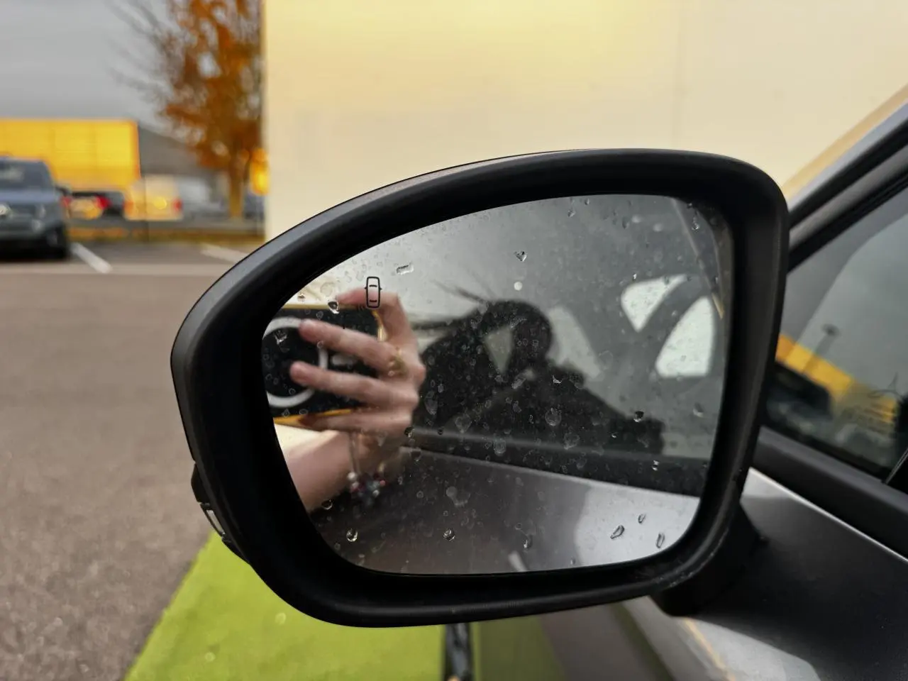 Rétroviseur extérieur droit noir brillant de Renault Clio gris schiste, avec traces d'eau et reflet de main tenant un appareil photo.