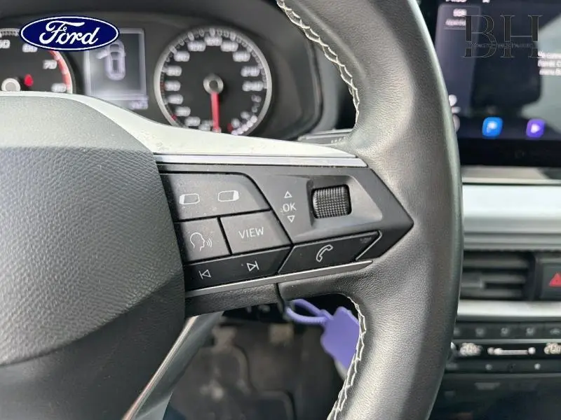 Gros plan sur les commandes au volant cuir noir du SEAT ARONA 2022, avec tableau de bord et écran tactile en arrière-plan.