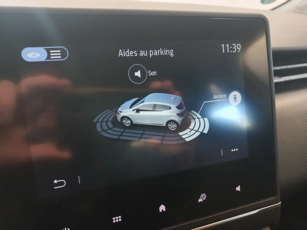 Affichage tactile intérieur montrant l'aide au parking avec vue 3D d'une Renault Clio blanche de profil côté gauche.