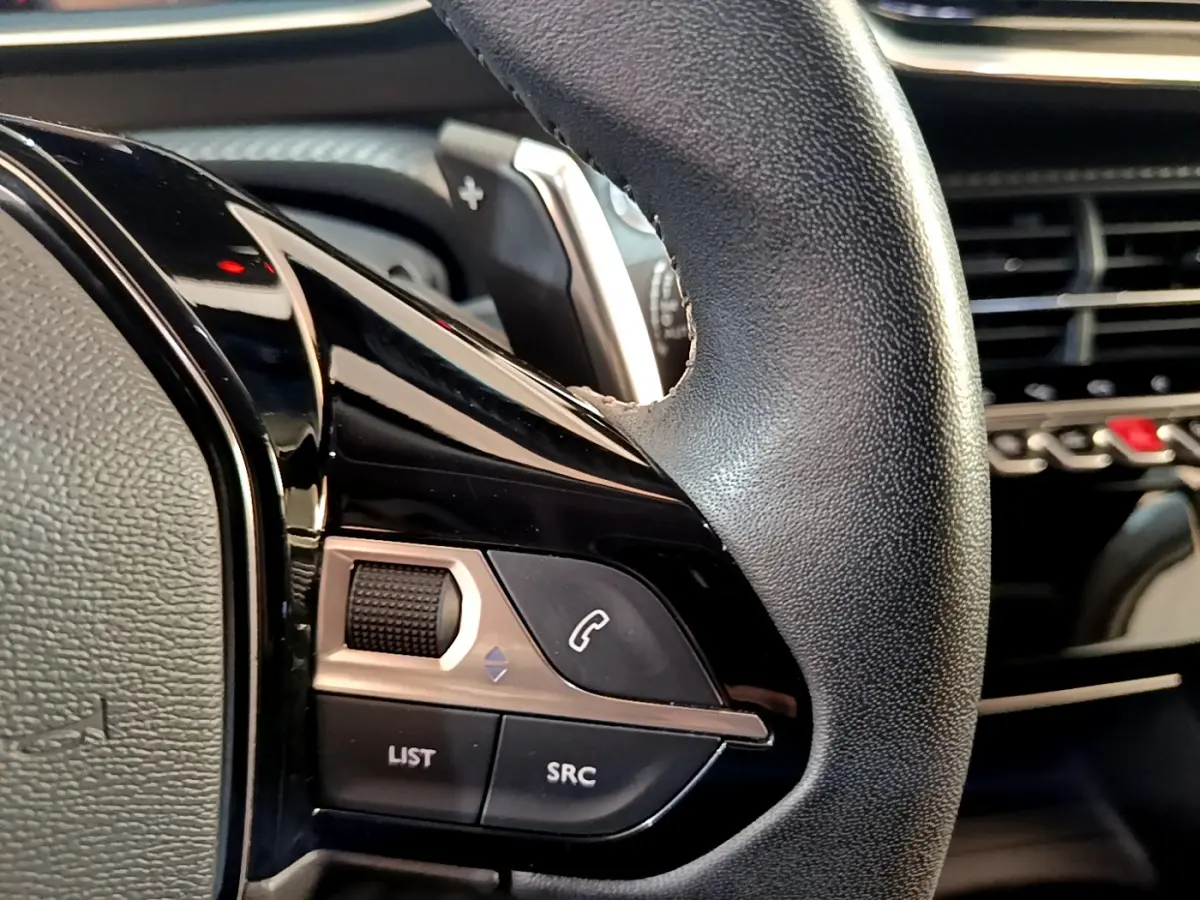 Gros plan sur les commandes gauche du volant cuir du Peugeot 2008 gris clair, avec tableau de bord en arrière-plan.