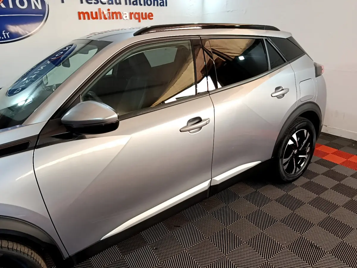Profil côté gauche d'un Peugeot 2008 gris clair 2021 avec jantes noires et toit noir dans un showroom.