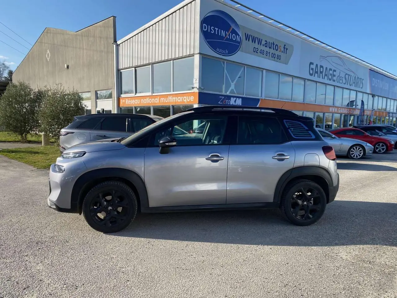 Vue de profil côté gauche d'un Citroën C3 Aircross gris foncé 2024 avec jantes noires, stationné devant un garage.