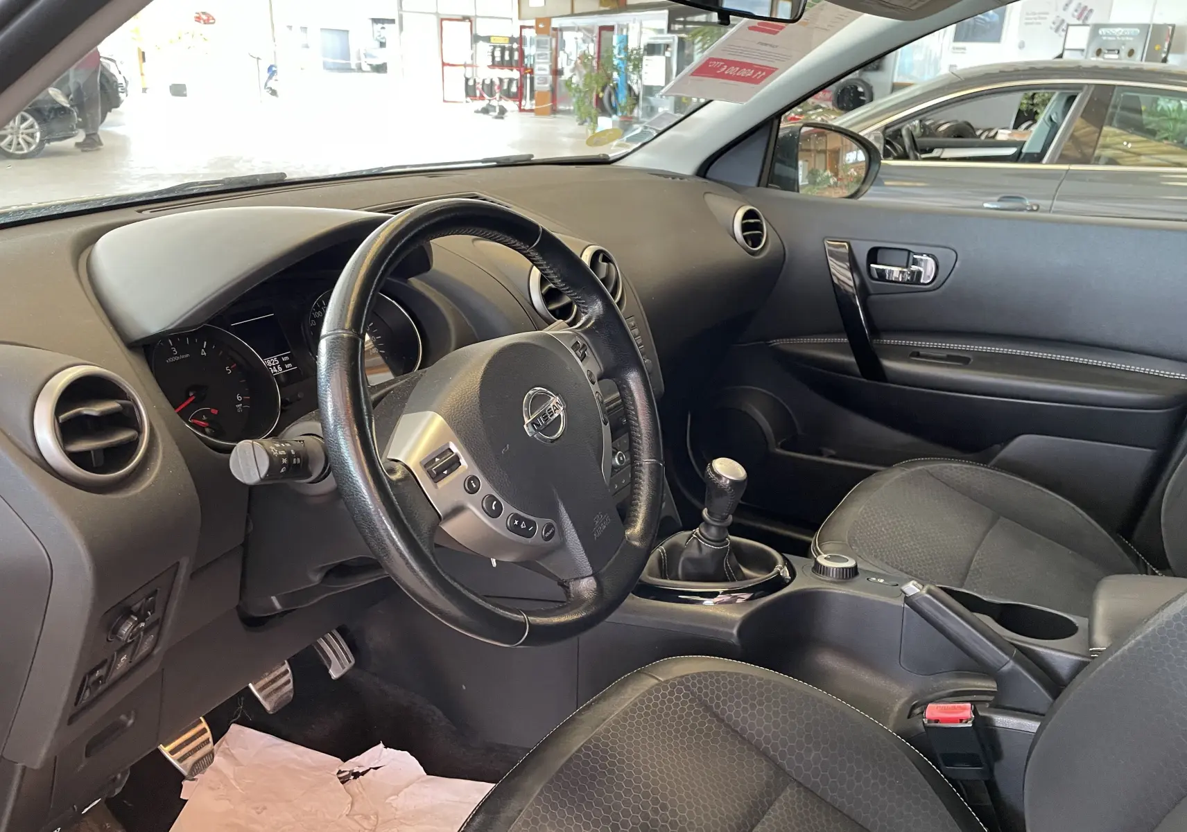 Intérieur noir du Nissan Qashqai 2013 vu côté conducteur, volant cuir, levier de vitesse manuel et tableau de bord visible.