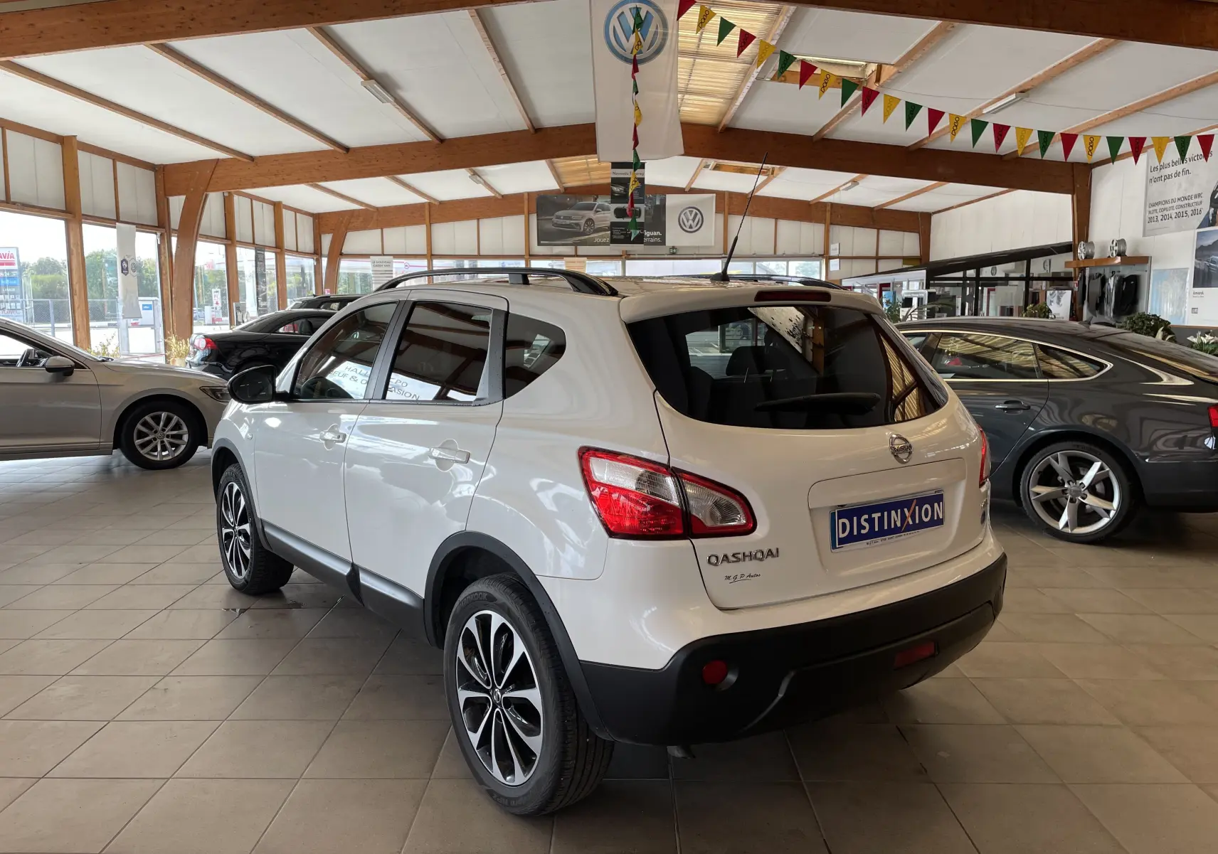 Vue 3/4 arrière droite d'un Nissan Qashqai blanc 2013 en showroom, avec jantes alliage et toit ouvrant visible.