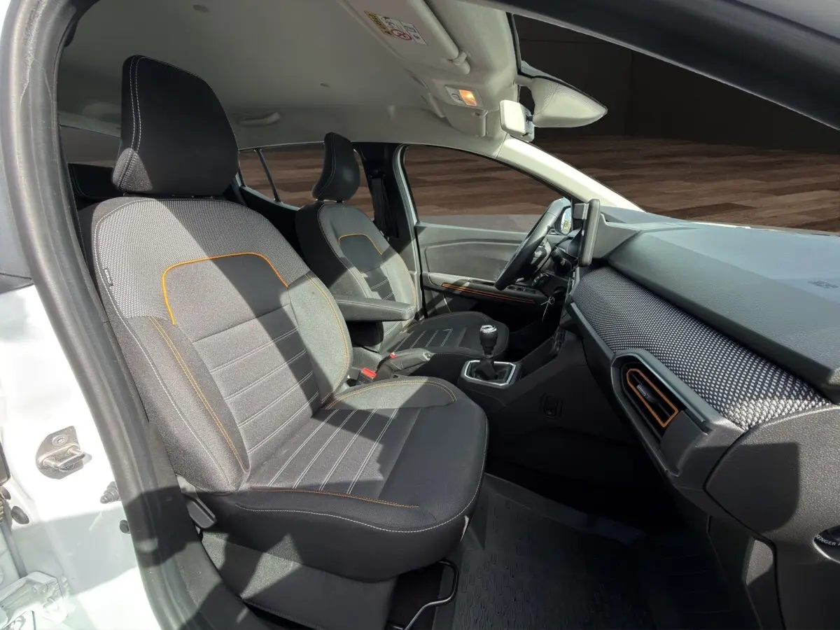Vue intérieure côté conducteur de la Dacia Sandero Stepway blanche, sièges tissu gris avec surpiqûres orange et tableau de bord moderne.