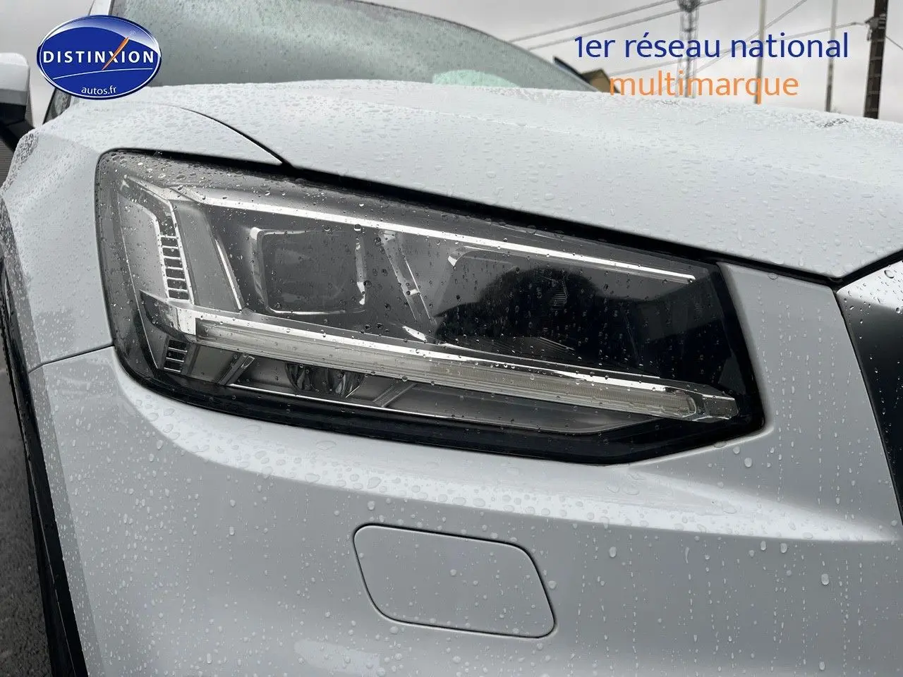 Gros plan sur le phare avant gauche d'un Audi Q2 blanc Ibis sous la pluie, mettant en valeur son design LED.