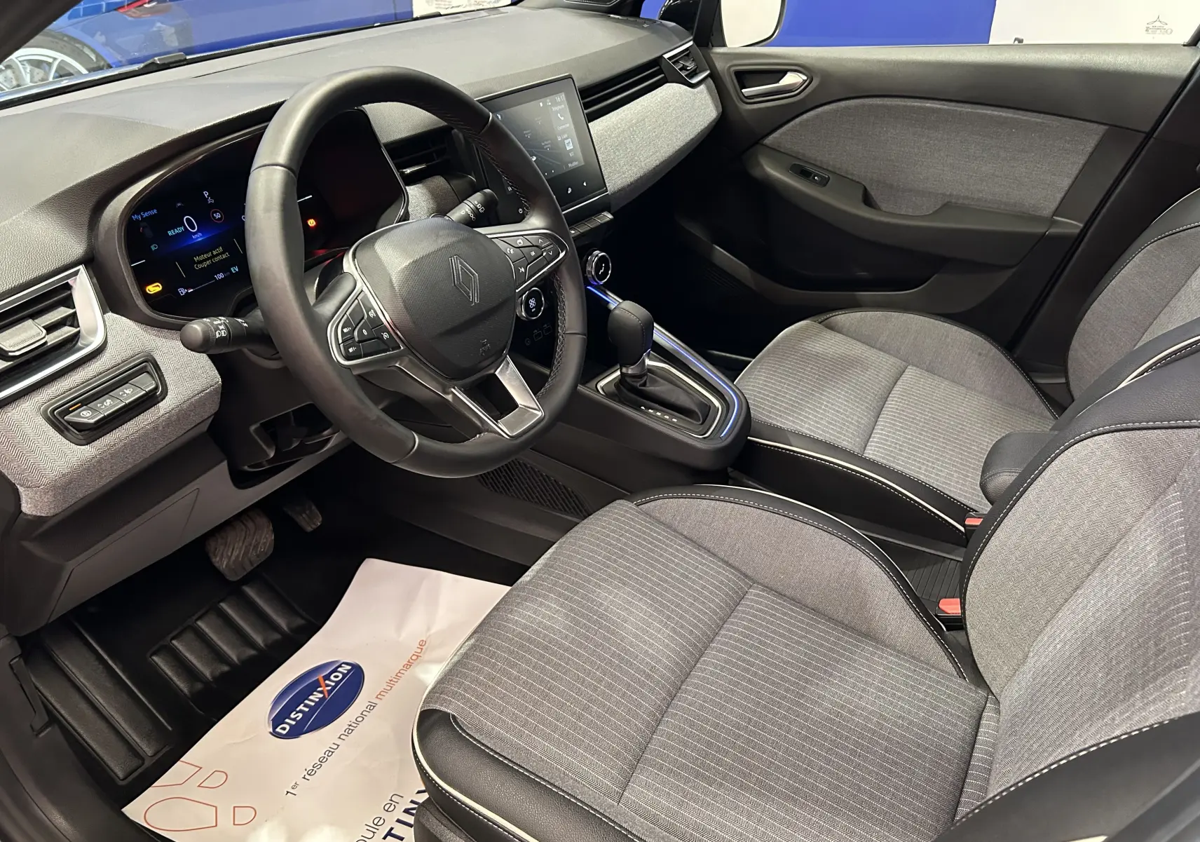 Vue intérieure côté conducteur de la Renault Clio V hybride, avec sièges gris tissu et tableau de bord moderne.