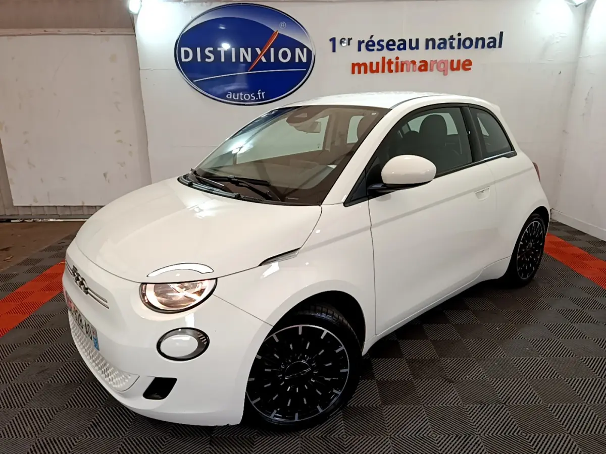 Fiat 500 électrique blanc vue 3/4 avant droit en intérieur avec jantes noires et phares allumés.