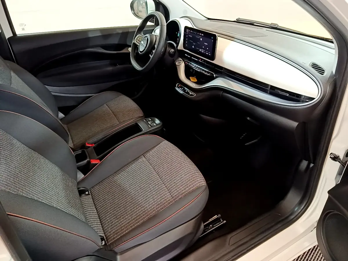 Intérieur avant droit de la Fiat 500 électrique blanche 2022, sièges tissu gris et tableau de bord avec écran tactile central.