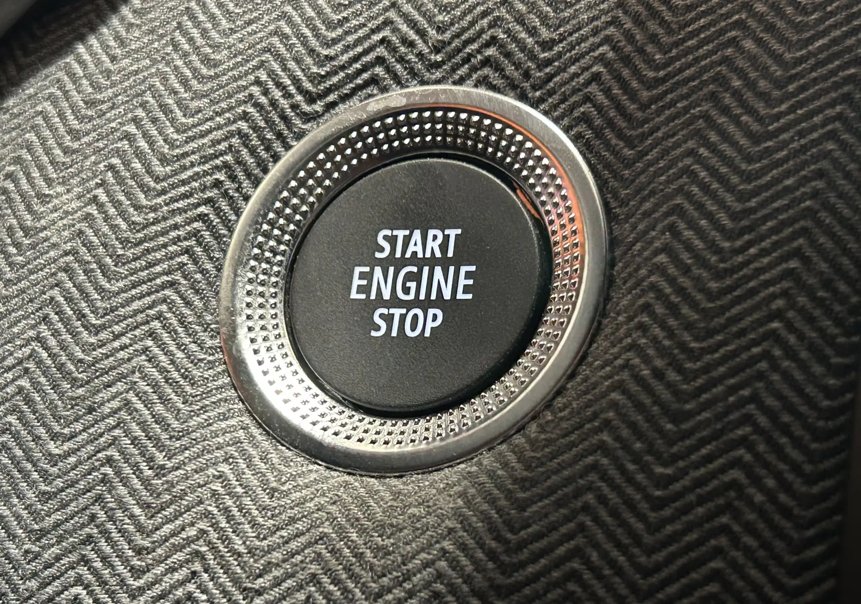 Gros plan sur le bouton Start Engine Stop entouré d'un cerclage métallique, sur un tableau de bord à motif chevrons gris.