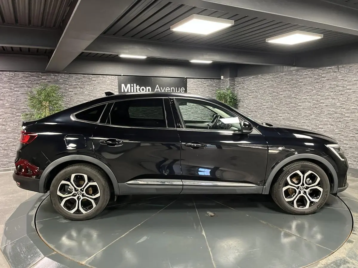 Profil côté gauche du Renault Arkana noir métallisé 2021 avec jantes alliage et lignes fluides dans un showroom intérieur.