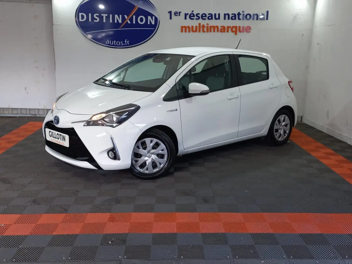 Toyota Yaris blanche vue de profil côté gauche en intérieur, avec phares allumés et logo hybride visible sur l'aile avant.