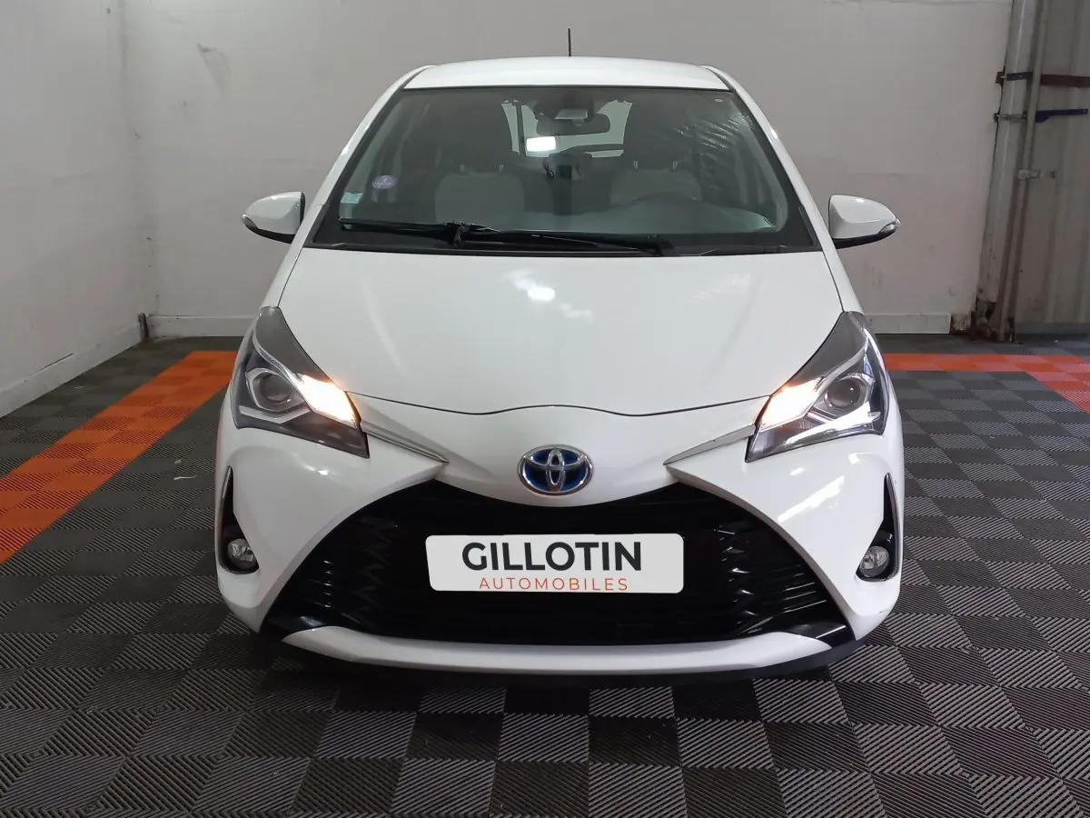 Vue frontale d'une Toyota Yaris blanche 1.5 VVT-I Hybrid avec phares allumés dans un garage.
