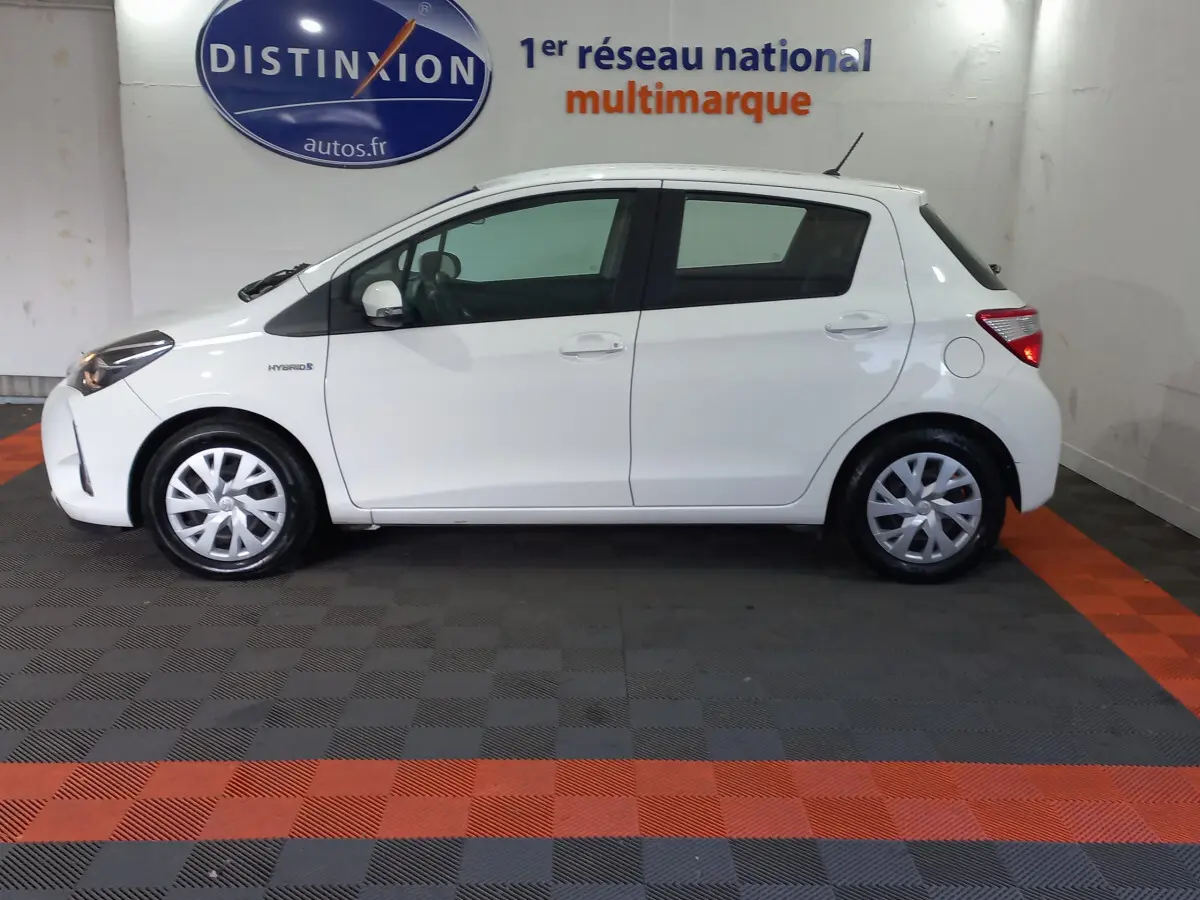 Profil gauche d'une Toyota Yaris blanche 2019 hybride, vue en intérieur de showroom avec logo Distinxion en arrière-plan.