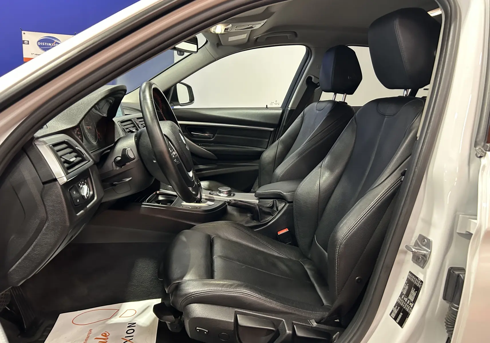 Vue intérieure côté conducteur de la BMW Série 3 320i blanche avec sièges cuir noirs et volant multifonction.