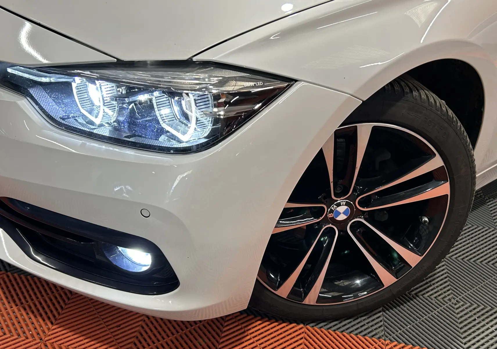 Gros plan sur l'avant droit blanc d'une BMW Série 3 320i 2018, mettant en valeur le phare LED et la jante bicolore.