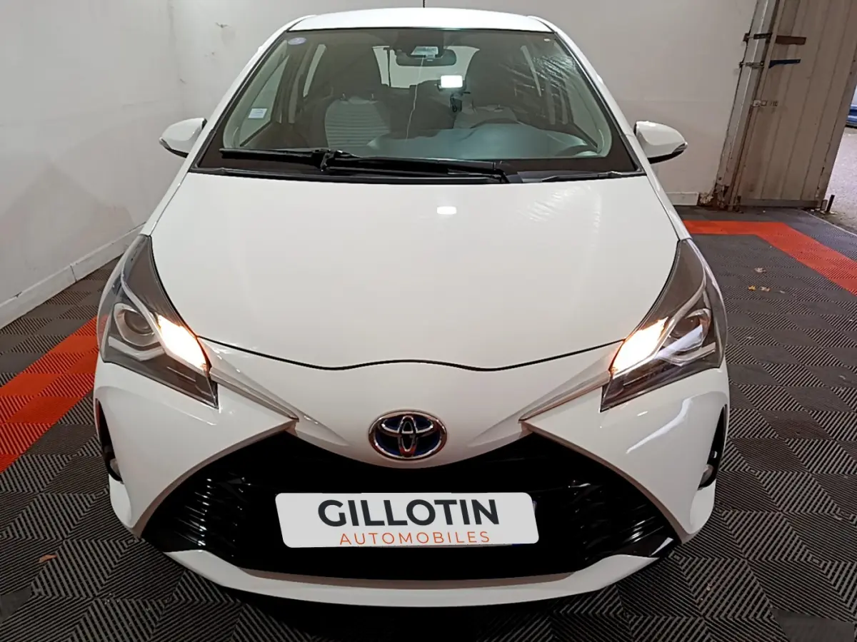 Toyota Yaris blanche vue de face avec phares allumés et calandre noire distinctive dans un garage.