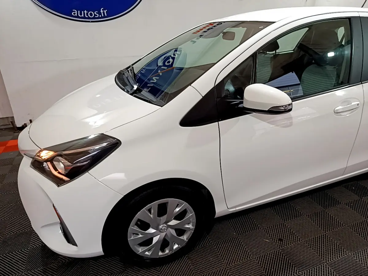 Toyota Yaris blanche vue 3/4 avant droit, phare allumé et rétroviseur avec clignotant intégré.