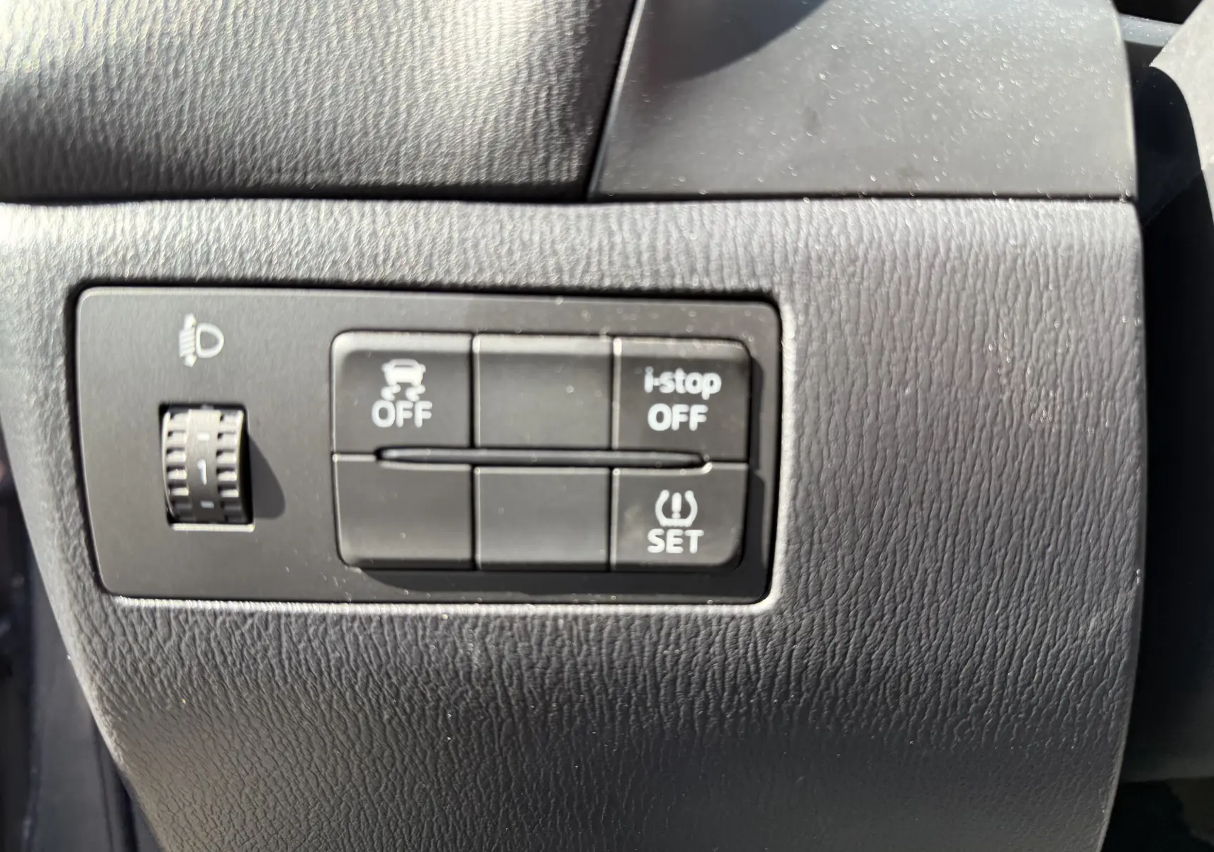 Gros plan sur les boutons de commande noirs du tableau de bord de la Mazda3 gris, avec réglage des phares et fonctions i-stop et contrôle de stabilité.