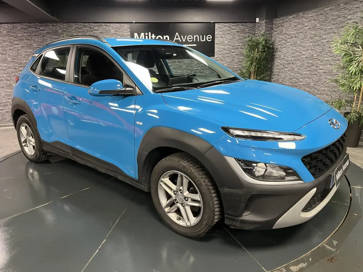 Hyundai Kona bleu 3/4 avant droit avec protections noires et jantes alliage 16 pouces en intérieur showroom.