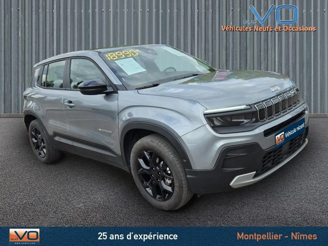 Jeep Avenger gris foncé en 3/4 avant droit, avec jantes noires et calandre distinctive, stationné devant un mur métallique.