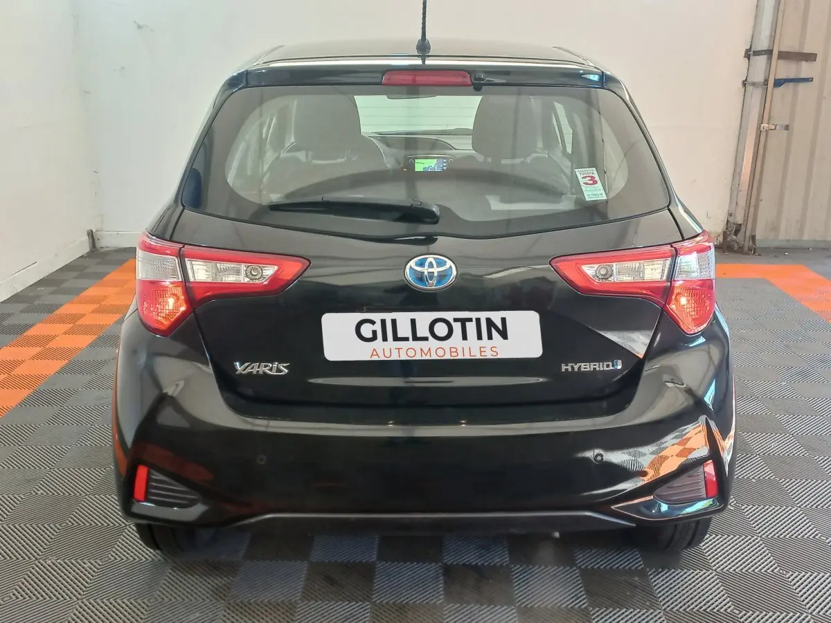 Vue arrière d'une Toyota Yaris hybride noire avec feux arrière allumés et logo visible sur un sol à damiers noir et orange.