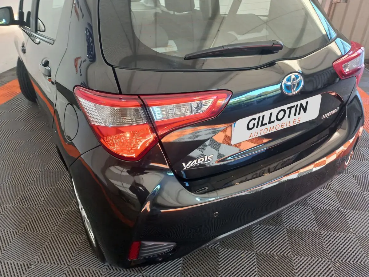 Vue 3/4 arrière gauche d'une Toyota Yaris noire hybride avec feux arrière distinctifs et logo visible.