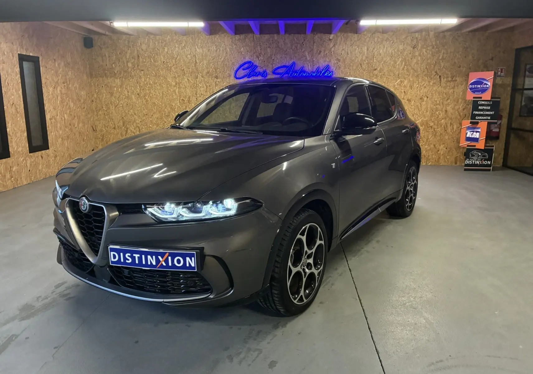 Alfa Romeo Tonale gris foncé vue 3/4 avant droit, phares allumés et jantes distinctives en intérieur showroom.