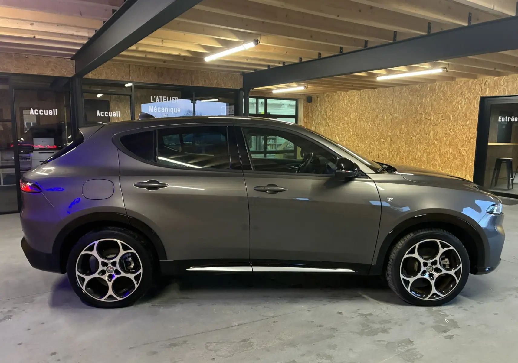 Vue de profil côté gauche d'un Alfa Romeo Tonale gris foncé 2023 en intérieur, avec jantes spécifiques et détails noirs.