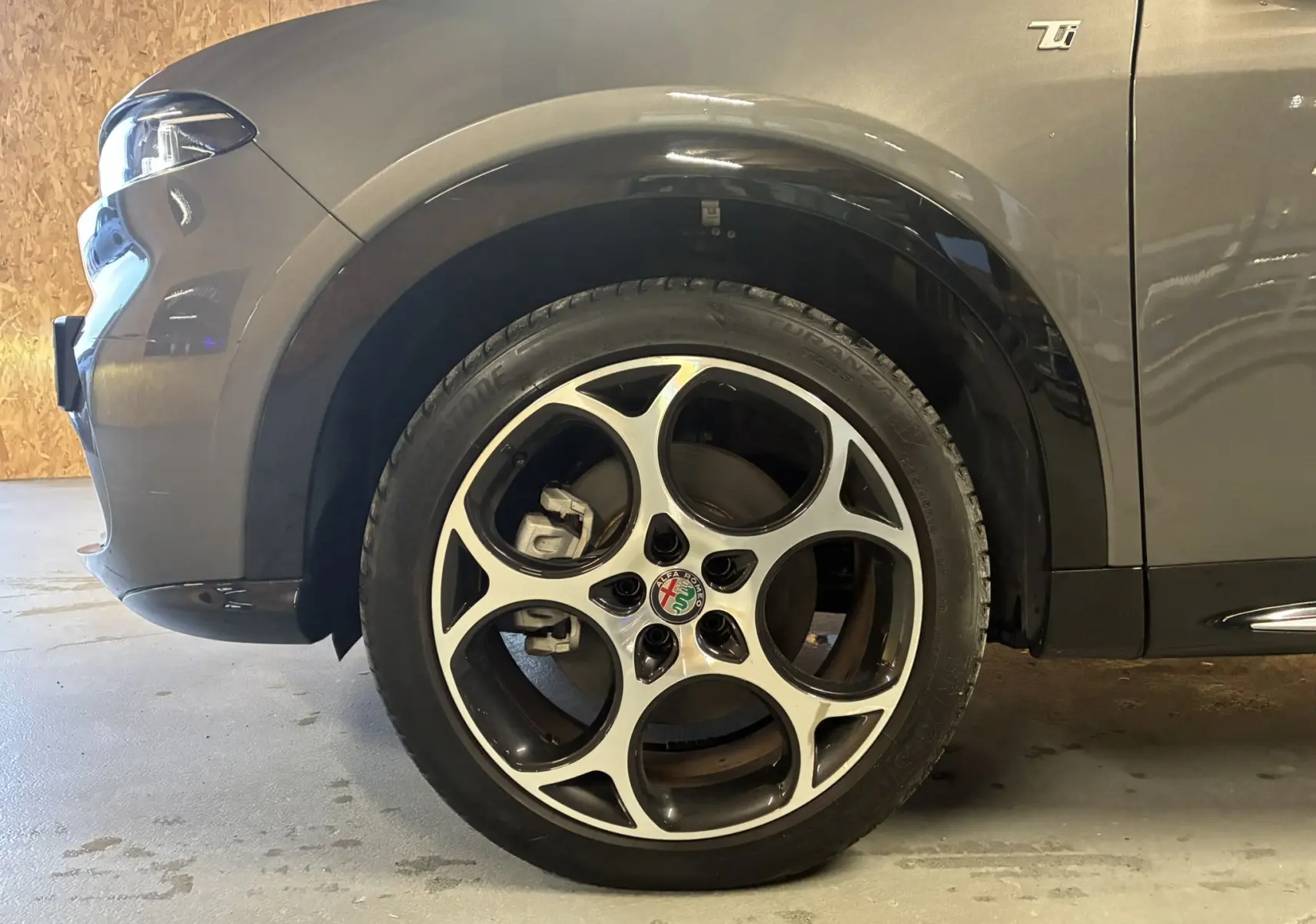 Gros plan sur la roue avant gauche et l'aile d'une Alfa Romeo Tonale gris foncé avec jante alliage distinctive à cinq cercles.
