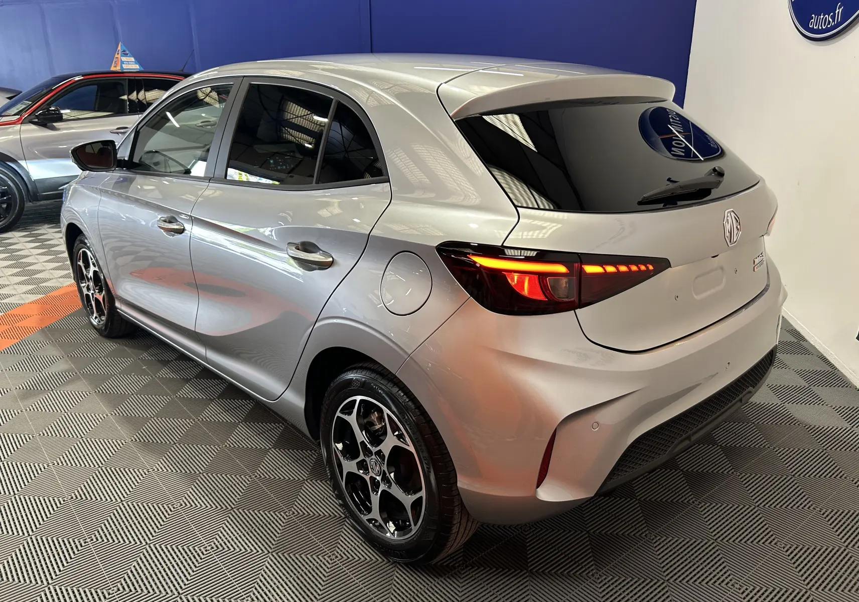 Vue 3/4 arrière droite de la MG3 1.5 Hybrid+ Cosmic Silver avec feux allumés et jantes noires brillantes.